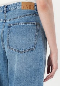 Primer plano del bolsillo de unos jeans de denim azul claro con una etiqueta de cuero marrón "VMD", mostrando la parte baja de la espalda y el lado del brazo y mano de una persona.