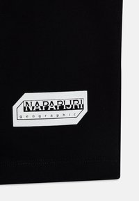 Svart tyg med en vit rektangulär etikett med texten "NAPAPURI geographic" i fet stil. Slät textur med sömmar på kanterna.