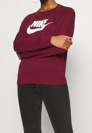 Persoon met gevouwen armen, gekleed in een bordeauxrode Nike-trui met wit logo en zwarte jeans tegen een effen lichte achtergrond.
