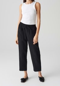 Pantalon noir ample avec une ceinture élastique et un cordon de serrage, doté de poches latérales et d'une coupe droite. Porté avec un débardeur blanc.