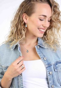 Zilveren sieraden set met turquoise-kleurige stenen; bevat druppelvormige oorbellen, een bijpassende hanger en een ring, uitgestald tegen een spijkerjack.