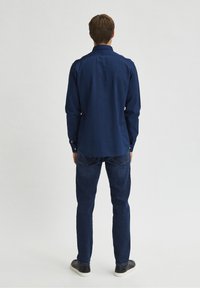 Muž čelící zády, oblečený do tmavě modré košile s dlouhým rukávem, modrých džínů a černých tenisek s bílými podrážkami na jednobarevném pozadí.