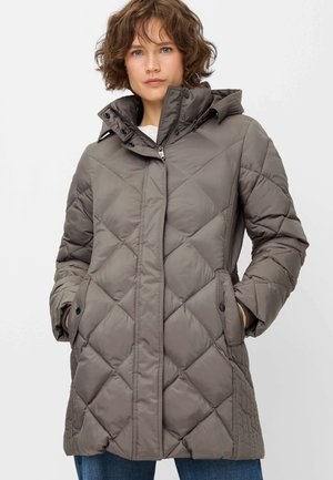 Woman wearing a knee-length taupe quilted puffer coat with a high collar, front zipper, snap pockets, and hands in pockets.