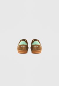 Barna színű, velúr sportcipő mentazöld díszítéssel, gumitalppal és hímzett márkajelzéssel a sarkon és az oldalak mentén. Texturált hátoldali kialakítás.