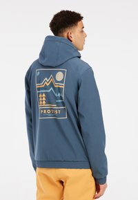 Blaue Kapuzenjacke mit einem grafischen Design, das Berge, Bäume und eine Sonne zeigt. Gerippte Ärmelbündchen und Saum mit Logo auf der Rückseite.