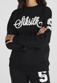 Czarna bluza z białym haftem "Siksilk" z przodu oraz białą łatką z numerem 5 na prawym rękawie. Miękka, luźna forma.