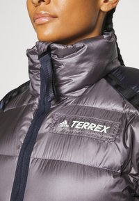 Puszysta szara kurtka z wysokim kołnierzem, ciemnoniebieskimi akcentami i białym logo adidas TERREX na piersi. Gładka, błyszcząca faktura materiału.