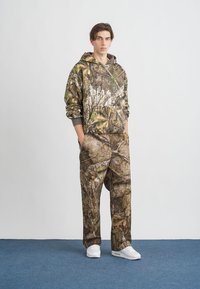 Kamuflázs pulóver és nadrág szett barna és zöld árnyalatokban, természetes mintával. A modellen fehér sportcipő van, és a frontzsebeit mutatja.