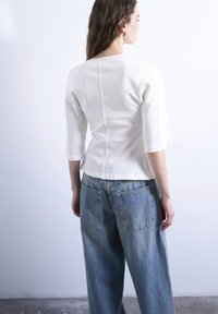 Γυναίκα με κυματιστά καστανά μαλλιά στέκεται προς τα πίσω, φορώντας λευκό μακρυμάνικο μπλουζάκι και άνετο μπλε τζιν με πίσω τσέπες.