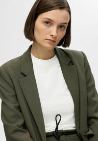 Olijfgroene oversized blazer van textuurstof, gecombineerd met een witte ronde hals t-shirt. Zwart trekkoord detail bij de taille.