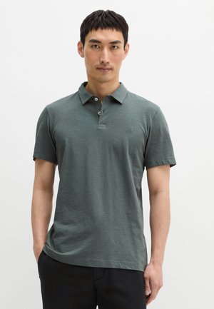 Mężczyzna w zielonej koszulce polo z krótkim rękawem i czarnych spodniach, stojący z rękami w kieszeniach na jednolitym białym tle.