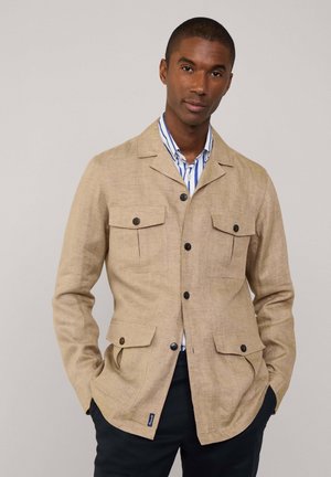 Beige linnen jas met een scherpe kraag, vier voorzakken en donkere knopen. Draagt over een blauw-wit gestreept shirt.