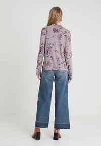 Långärmad lavendelfärgad topp med blommönster och fågelmotiv, i kombination med vida jeansbyxor med fransade kanter och bruna klackskor.