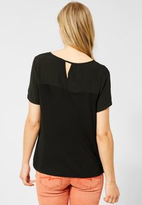 Frau mit langen blonden Haaren, die ein schwarzes kurzärmliges Oberteil mit einem Rückenausschnitt und korallfarbene Hosen trägt, steht vor einem einfarbigen Hintergrund.