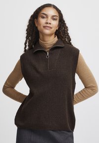 Brun ribbet ærmeløs vest med høj hals og lynlås halvvejs. Bæres over en langærmet tan skjorte. Minimalistisk design.