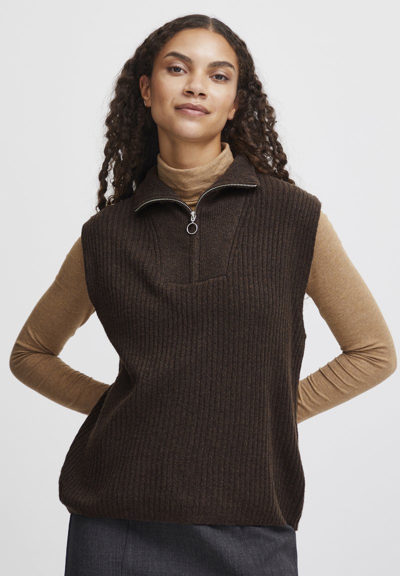 Brun ribbet ærmeløs vest med høj hals og lynlås halvvejs. Bæres over en langærmet tan skjorte. Minimalistisk design.