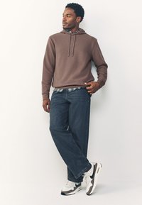 Felpa con cappuccio marrone con trama, con coulisse, indossata sopra una camicia a quadri, abbinata a jeans blu a taglio ampio e sneakers bianche con dettagli grigi.