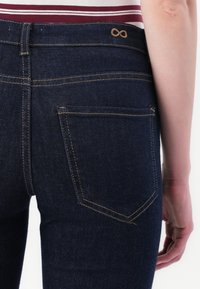 Κοντινή λήψη ενός ατόμου που φορά σκούρα μπλε τζιν παντελόνι με καφέ ραφές και ένα κεντημένο σύμβολο άπειρου πάνω από την πίσω τσέπη.