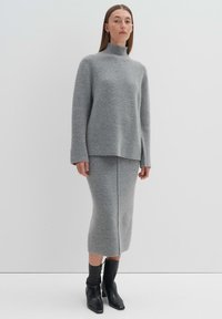 Helle graue Strickjacke mit hohem Kragen, langen Ärmeln, kombiniert mit einem passenden knielangen Rock. Das Model trägt schwarze Knöchelstiefel.