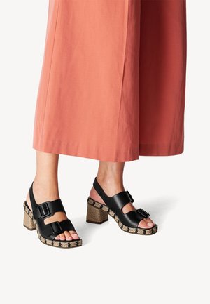 Sorte sandaler med hæl og to stropper med spænder, flettet sål med jute-lignende tekstur og blokhæl. Sammen med et koralfarvet bukseben med vide ben.