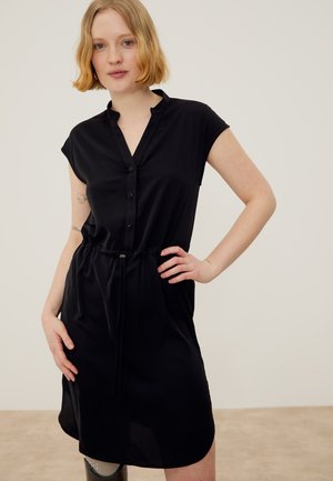 Γυναίκα με κοντά ξανθά μαλλιά, που φοράει μαύρο φόρεμα με κουμπιά, κοντά μανίκια και ελαφρώς ψηλή μέση, στέκεται με το ένα χέρι στη μέση της.