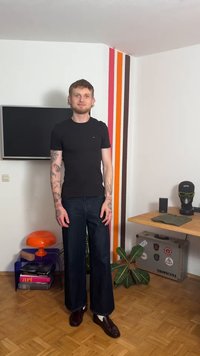 Schwarzes T-Shirt mit kurzen Ärmeln, blaue weit geschnittene Jeans, dunkelbraune Loafers. Auffällige Tattoos auf den Armen, stehend vor einer farbig gestreiften Wand.