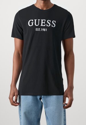 Camiseta de algodón negra con cuello redondo y mangas cortas, con el texto en plata "GUESS" y el año de fundación impresos en la parte delantera.