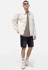 Young man wearing beige denim jacket, brown t-shirt, black denim shorts, white high-top sneakers, striped socks, and tinted sunglasses standing on white background.