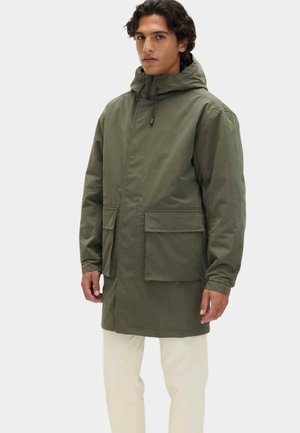 Mand iført olivengrøn hætteparka med store forlommer og beige bukser, stående mod en ensfarvet lys baggrund.