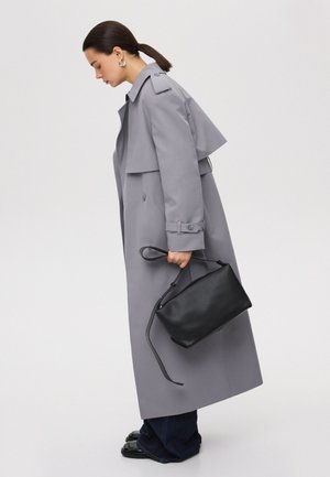 Grauer Trenchcoat mit strukturiertem Design, tailliertem Bund und übergroßen Revers; kombiniert mit einer schwarzen Lederhandtasche und dunklen Jeans.