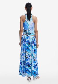 Rochie maxi cu gât halter, imprimeu floral vibrant albastru, fustă fluidă și textură de material neted. O pereche de sandale albe cu bretele.