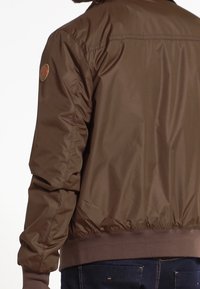 Veste bomber marron à texture lisse, poignets et ourlet côtelés, avec un écusson logo circulaire sur l'épaule gauche.