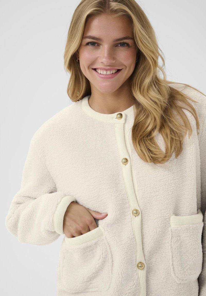 Young woman smiling, wearing a cream-colored textured cardigan with gold buttons, hands in front pockets, blonde hair over shoulder.