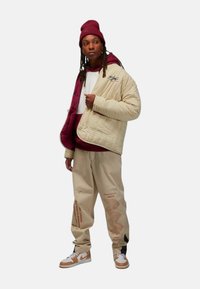 Beige quiltet jakke, burgundy hættetrøje, beige bukser med tekst, hvide sneakers med orange accenter, og matchende hue. Teksturede stoffer præsenteret.