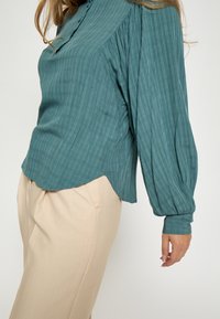 Tyrkysová textura blůza s kulatým výstřihem, sklady a dlouhými puff rukávy, v kombinaci s světle béžovými kalhotami.