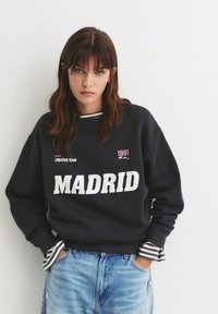 Čierna mikina s nápisom "MADRID" veľkými bielymi písmenami, s malou ružovou grafikou a pruhovanou košeľou viditeľnou na nápletoch. Nosené s modrými džínsy.