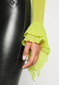 Transparent, genomskinlig långärmad topp i stark neon-gul med volangärmar; matchad med en slank, glansig svart läderkjol.