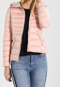 Jasnoróżowa kurtka puchowa z zapięciem na zamek z przodu, pikowany fason i kontrastowy, jasny kołnierz. Dwie kieszenie boczne z zamkami.
