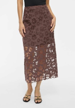 Femme portant un haut noir sans manches et une jupe midi en dentelle florale marron avec des talons ouverts noirs, debout devant un fond uni.