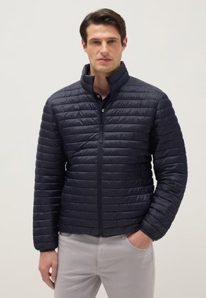 Mann trägt eine marineblaue gesteppte Jacke mit Reißverschluss und steht mit einer Hand in der Tasche einer hellgrauen Hose vor einem einfarbigen Hintergrund.