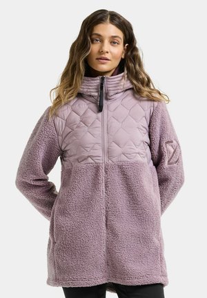 Femme aux cheveux ondulés portant une veste matelassée mauve en polaire avec un col montant et une fermeture éclair à l'avant, sur un fond uni.