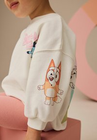 Sudadera blanca con personajes de dibujos animados coloridos en las mangas. Los puños son de canalé; la tela parece suave y con un ajuste relajado. Fondo claro.