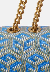 Blå tekstureret håndtaske hjørne med et guld geometrisk mønster og tykke guldkædestropper, der er trthreaded gennem guldøjer.