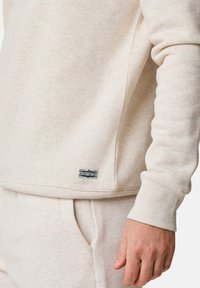 Beige sweatshirt with a textured fabric, ribbed cuffs, and a small logo tag on the hem. Simple design with a relaxed fit.