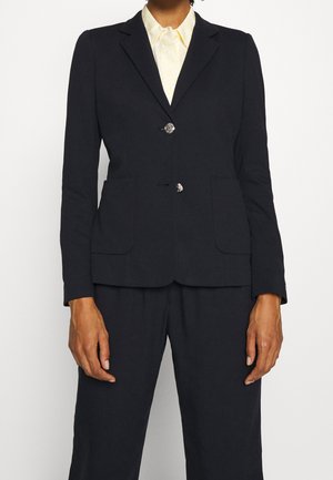 Persoon draagt een zwart colbert met twee knopen over een lichtgeel overhemd met kraag en bijpassende zwarte broek, staand tegen een effen achtergrond.