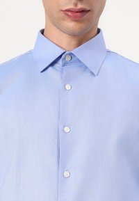 Gaiši zils krekls ar struktūru veidotu apkakli, ar baltām pogām un gludu, teksturētu audumu. Tuvplāns uz augšējo daļu.