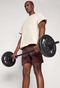 Chemise beige à manches courtes et shorts bordeaux portés par une personne tenant une plaque de haltère noire, mettant en valeur un design solide et des matériaux fonctionnels.