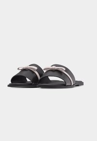 Paire de sandales slides noires avec des nœuds rose clair sur des larges brides, semelles plates et orteils ouverts sur fond blanc.