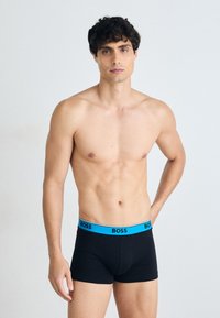 Schwarze Boxer-Shorts mit einem blauen Gummibund, der mit "BOSS" beschriftet ist. Hergestellt aus weichem, dehnbarem Material, mit einem figurbetonten Design, das den Körper umschmeichelt.
