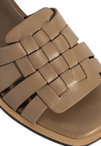 Sandale en cuir beige avec un design de lanière tissée, une texture lisse, une semelle plate et une forme de orteil arrondi.